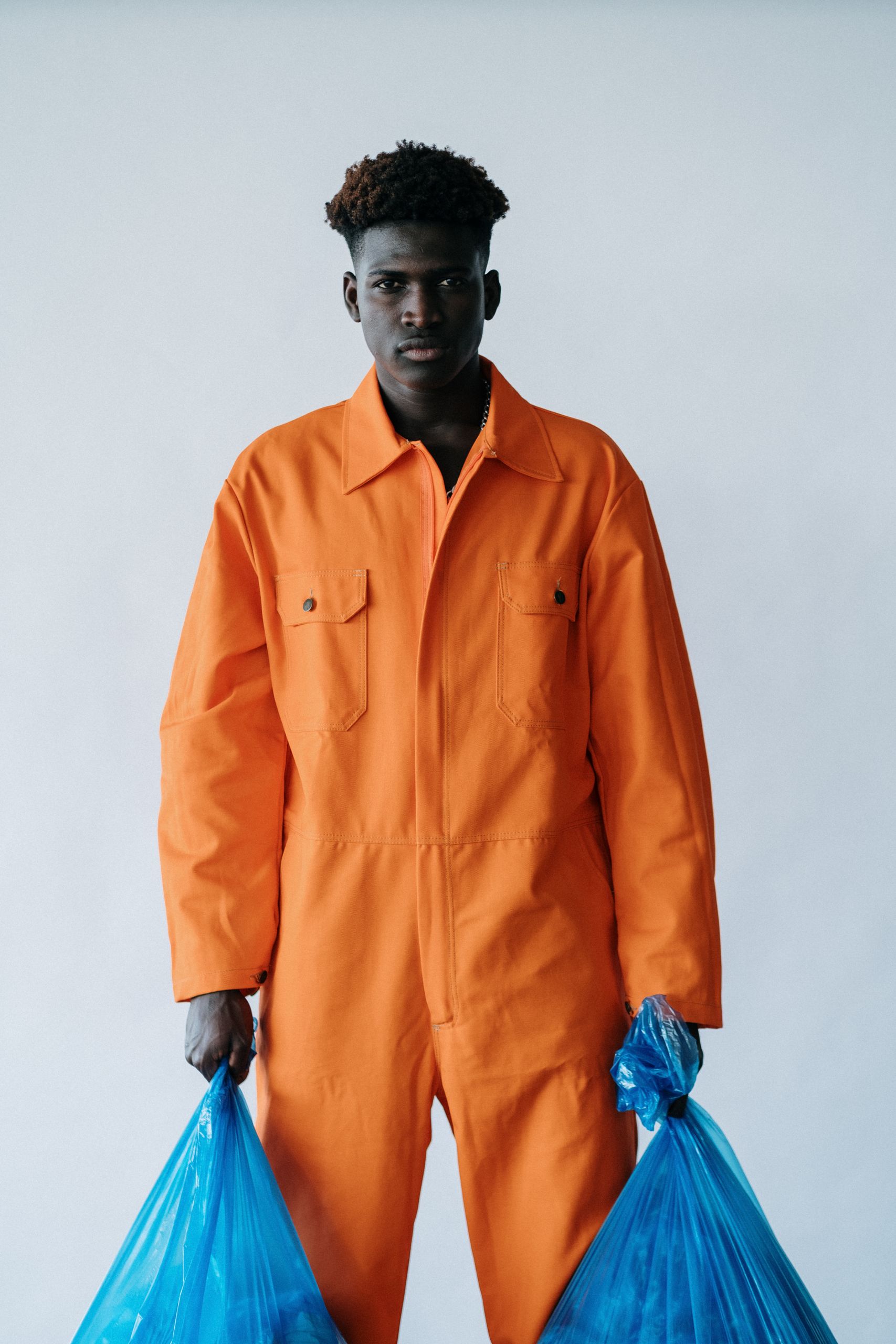 Black man in orange overalls holding blue garbage bags against a white background.